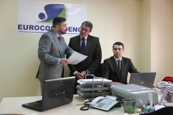 Thrivve by Intelletec | Your Next Job Hub Three men in suits gather around a desk with laptops, discussing ideas for Thrivve by Intelletec.