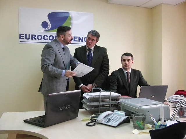 Three men in suits gather around a desk with laptops, discussing ideas for Thrivve by Intelletec.